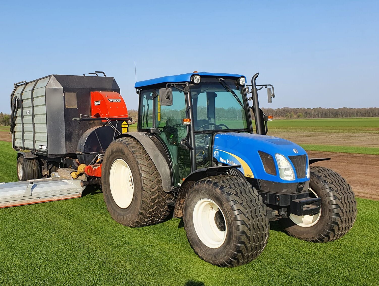 Een blauwe New Holland tractor met Trilo maai- en zuigcombinatie aan het werk op een strak groen graszodenveld.
