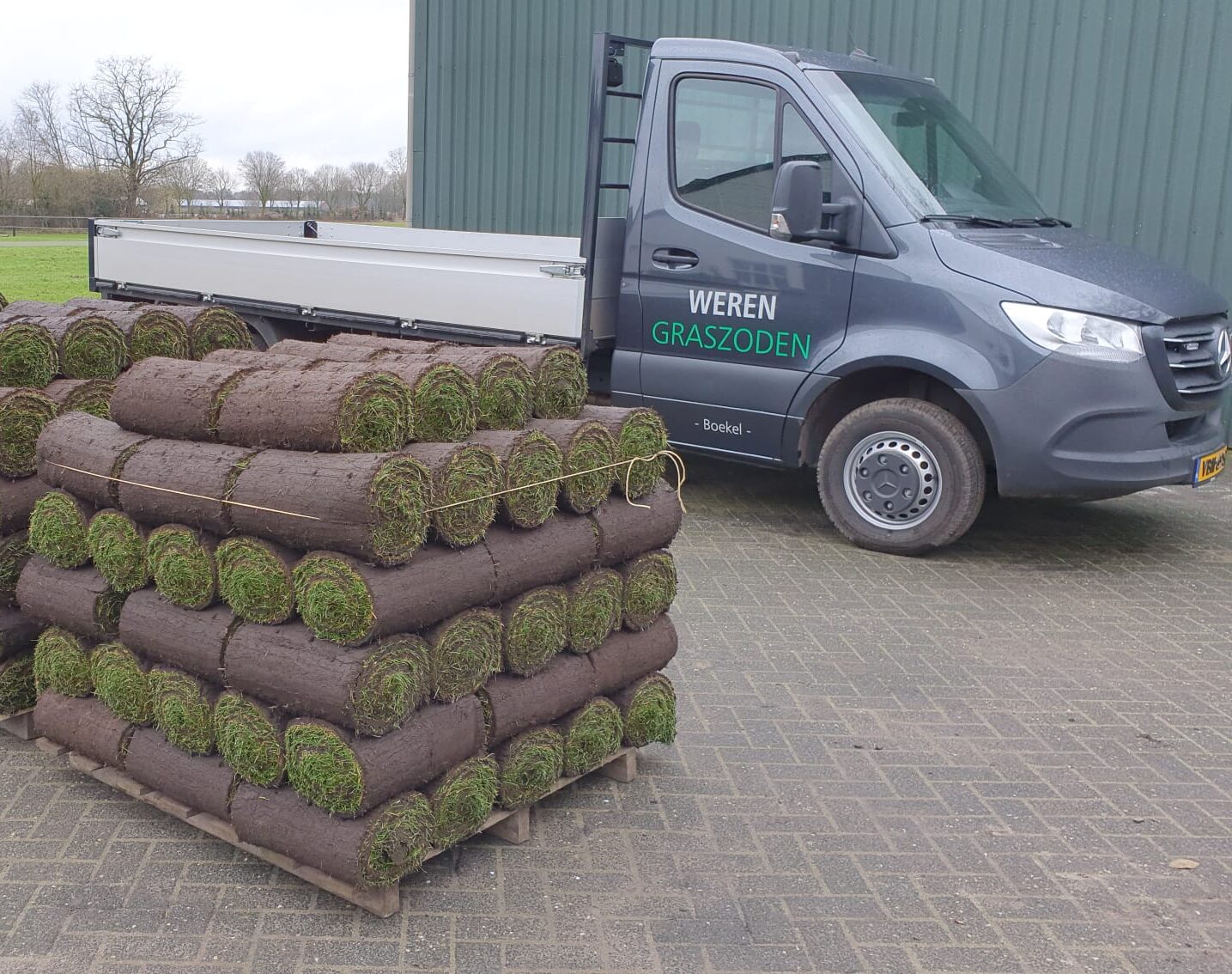 Pallets met vers gesneden rollen graszoden naast een grijze Weren Graszoden bedrijfswagen voor levering.